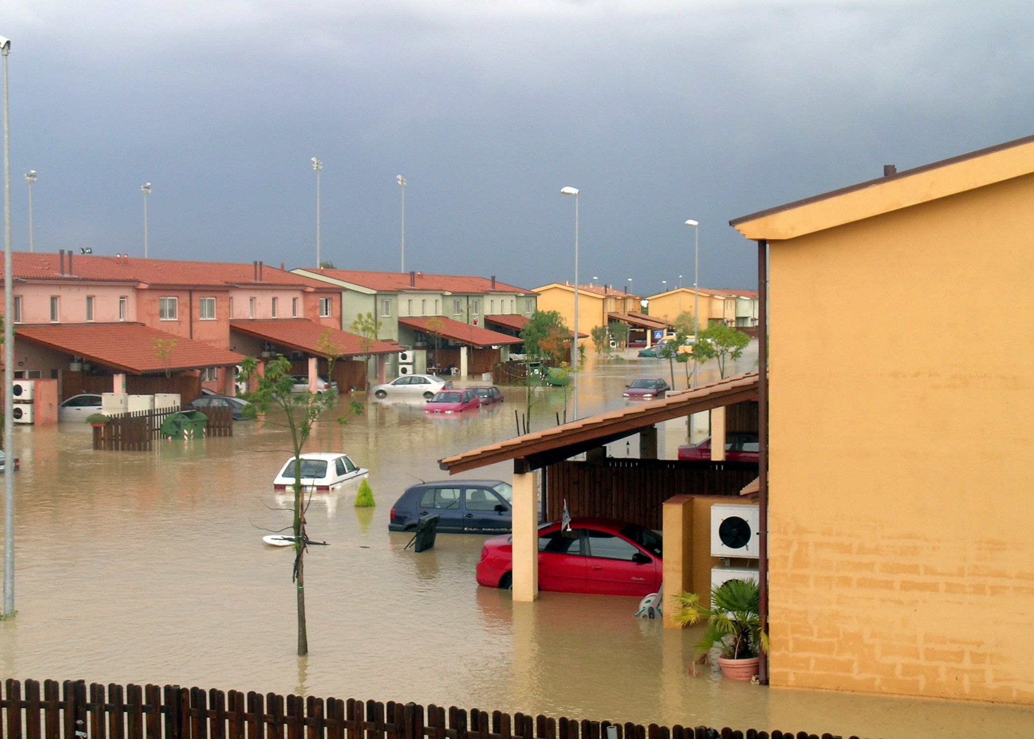 Immagine di una strada allagata tra due edifici, con acqua che copre l'intera superficie della strada. Sullo sfondo ci sono colline e nuvole grigie.