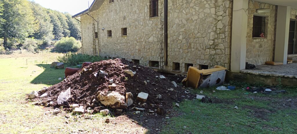 Un cumulo di terra e detriti abbandonati accanto a un edificio in pietra. Sono visibili anche rifiuti ingombranti, come divani e altri materiali, sul terreno erboso